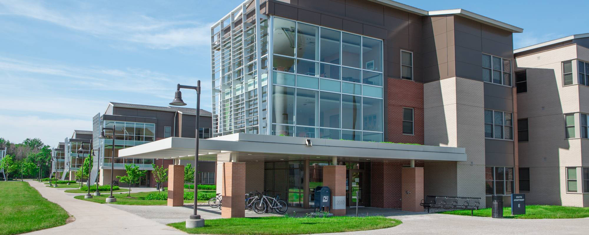 South Apartments building with large glass windows, brick accents, and bike racks. A clear blue sky and green lawn create a calm atmosphere.
